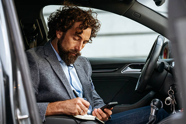 Focused businessman in blazer sits inside his vehicle, taking notes in a notebook.